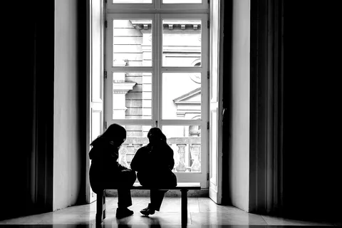 Due donne sedute di spalle davanti a una grande vetrata, ridotte a sagome dal controluce